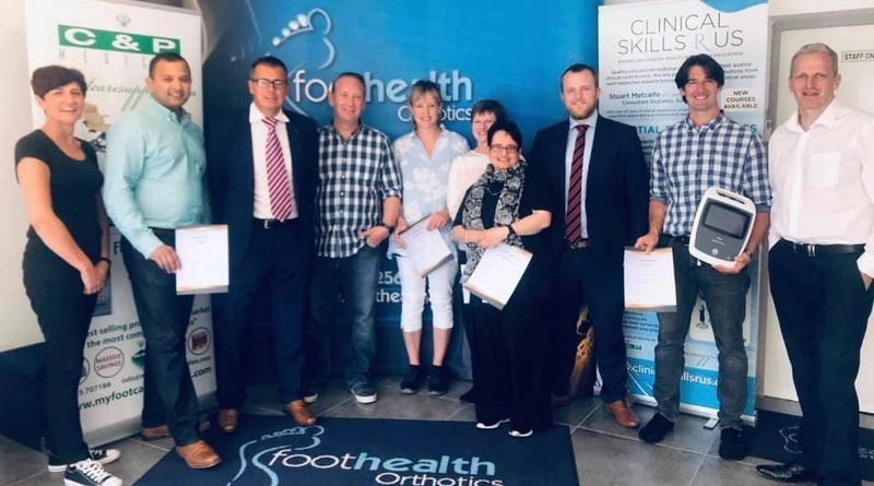 A group of professionals is standing together in front of banners for "Foot Health Orthotics" and "Clinical Skills R Us." The group consists of men and women dressed in business casual attire. Some individuals are holding certificates, indicating a formal event or training completion. The setting is a clinic or professional environment focused on foot health and orthotics.