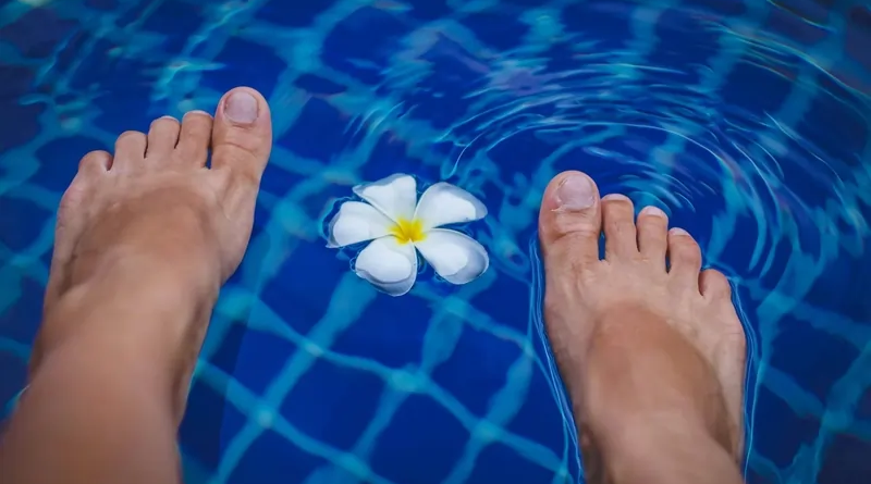 A pair of feet dipping into the clear blue water of a swimming pool with a yellow and white frangipani flower floating nearby. The water's surface shows a tiled pattern in shades of blue.