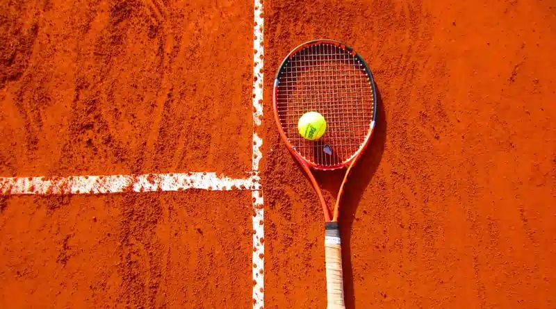 A tennis racket and a tennis ball are placed on a clay tennis court near the intersection of the white boundary lines. The image is taken from an overhead perspective.