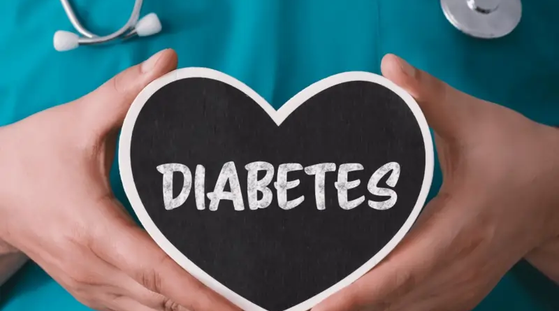 A person in a medical scrub top is holding a black heart-shaped sign with the word "DIABETES" written in white chalk. The person has a stethoscope around their neck.