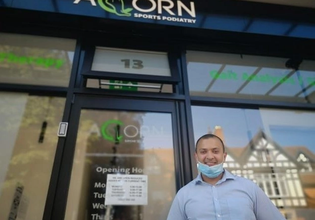 A man wearing a face mask below his chin stands in front of the entrance to Acorn Sports Podiatry. The glass door displays the clinic's name, logo, and opening hours. The man is smiling and dressed in a light blue shirt.