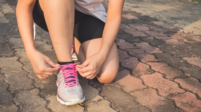 A person wearing a white shirt and black shorts is kneeling on a brick pathway, tying the pink laces of their gray running shoe.