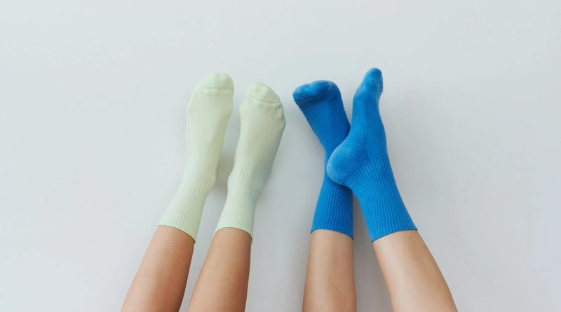 Two sets of feet wearing mismatched socks, one green pair and one blue pair, are extended upwards against a plain white background.