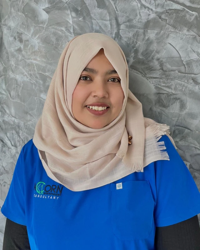 A smiling female healthcare professional wearing a light-colored headscarf and blue scrubs with 'ACORN Sports Podiatry' embroidered on the uniform, standing against a textured gray wall. She appears friendly and approachable.