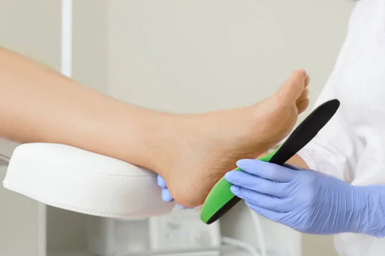 Home 4 A healthcare professional wearing blue gloves adjusts a green and black orthopedic insole under a patient's foot, which is resting on a cushioned support. The setting appears to be a medical or podiatry clinic.
