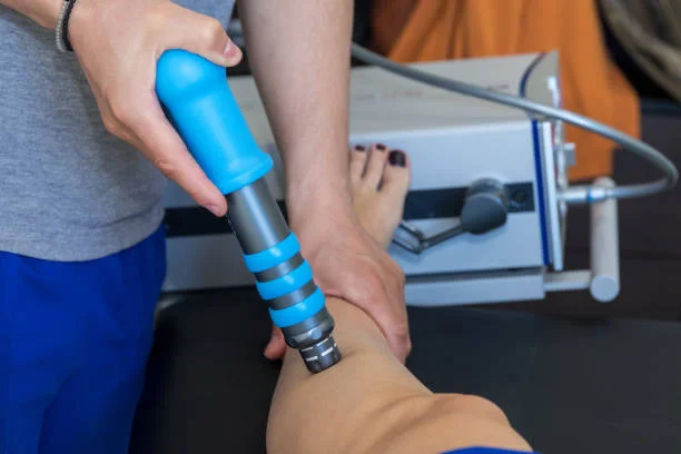 Home 5 A therapist is performing shockwave therapy on a patient's leg using a blue and gray device connected to a machine. The patient is lying down, and the focus is on the therapist's hand holding the device against the patient's leg.