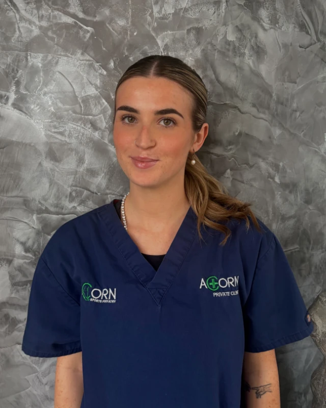 A smiling female healthcare professional in blue scrubs with "ACORN Sports Podiatry" embroidered on her uniform stands against a textured gray wall. She has brown hair pulled back and appears friendly and approachable.