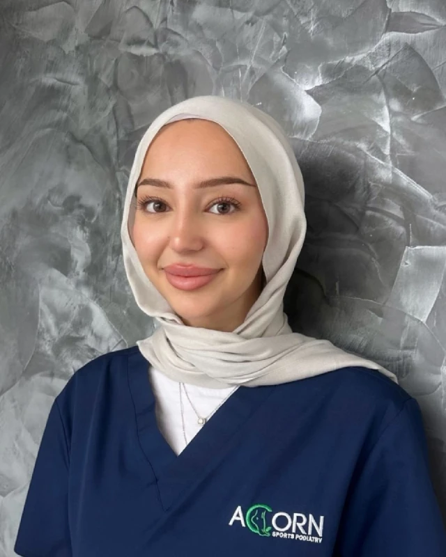 A smiling female healthcare professional wearing a light-colored headscarf and blue scrubs with 'ACORN Sports Podiatry' embroidered on the uniform, standing against a textured gray wall. She appears friendly and approachable.